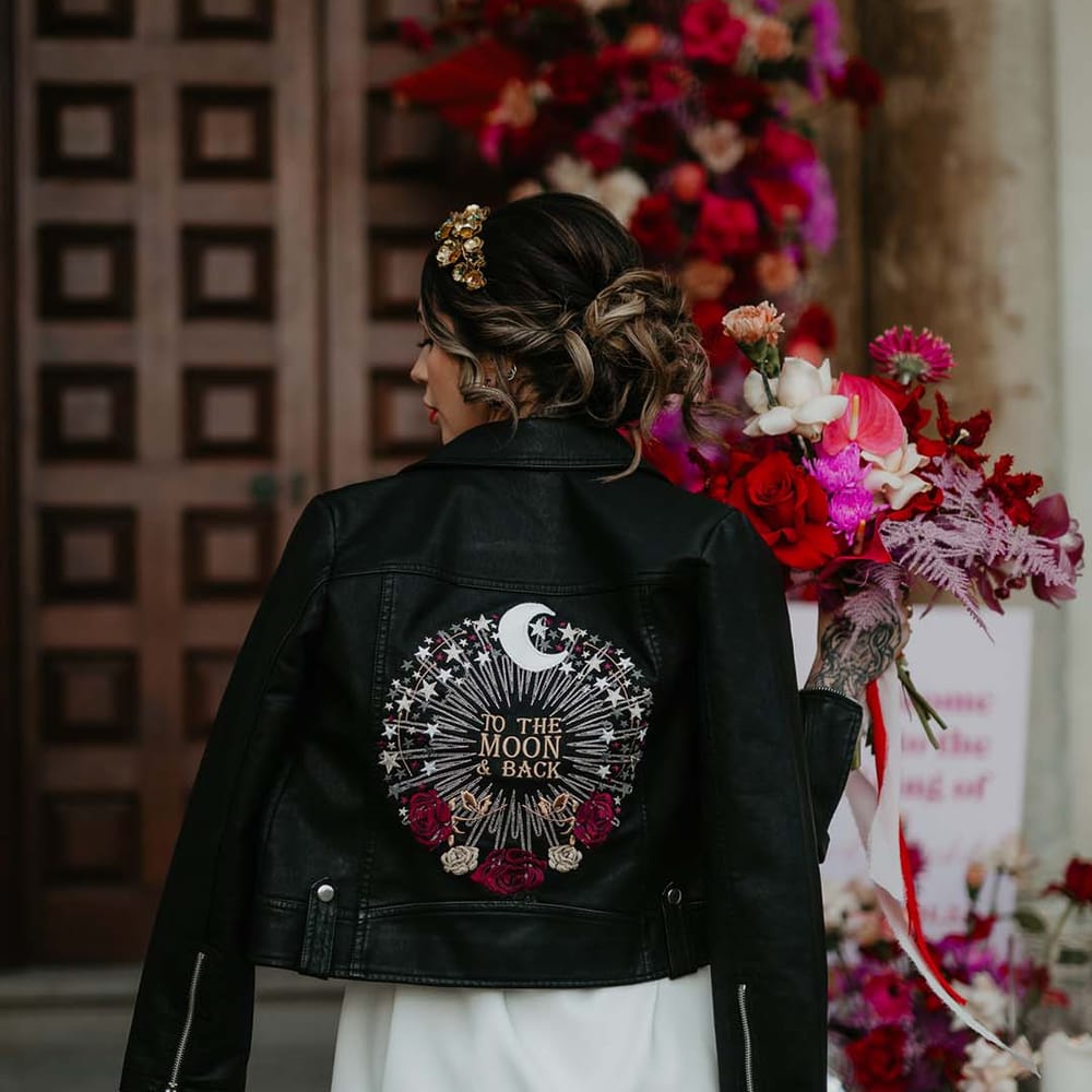 Bride in blazer style wrap around short wedding dress with delicate fringe detailing and black bridal leather jacket with “to the moon and back” lettering with crescent moon, stars, and roses embroidery 