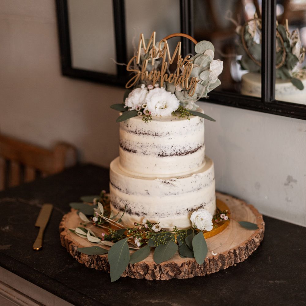 White frosted naked wedding cake complete with floral decor and gold personalised cake topper