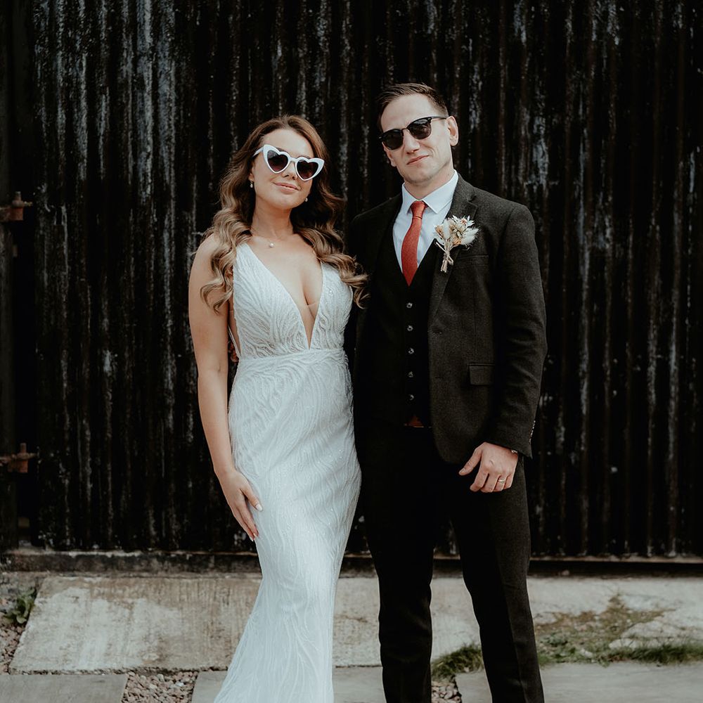 The bride in a sequin embellished Made With Love wedding dress and white retro sunglasses with the groom wearing black sunglasses to match the black suit 