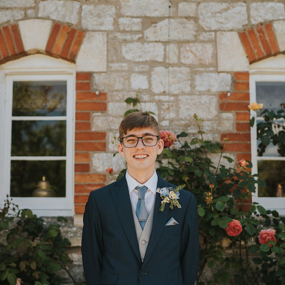 The groom in a navy suit jacket with light grey waistcoat with light blue tie and daisy buttonhole with glasses 