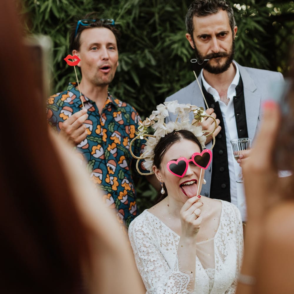 Bride in white bridal crown holding loveheart sunglasses prop with guests at wedding photobooth
