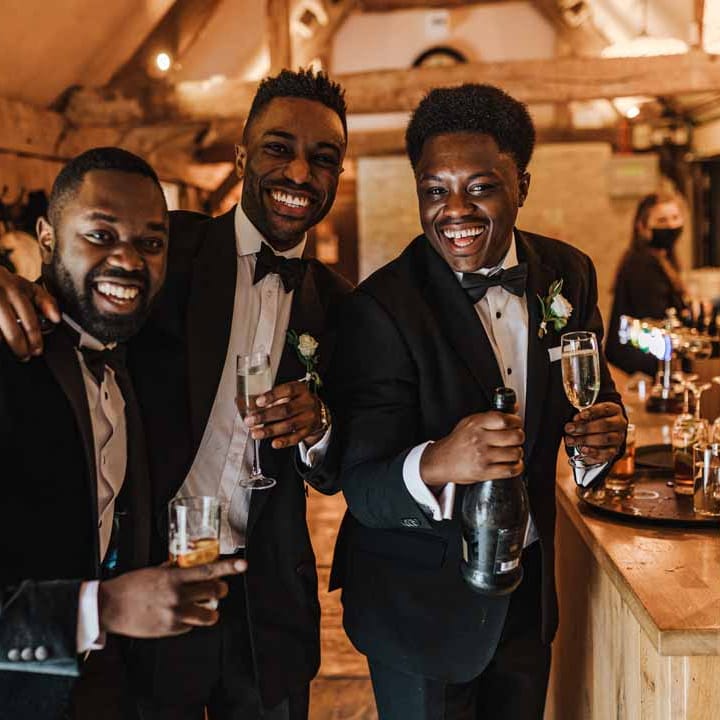 Groomsmen wearing classic black suits with bowties and white garden rose and foliage boutonnieres holding glasses of champagne and smiling at Irish Ghanaian fusion wedding