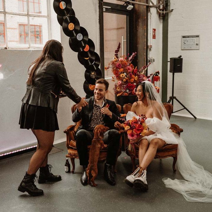 Wedding celebrant wearing leather jacket greeting bride and groom sat by vinyl wedding arch in crushed velvet armchairs 