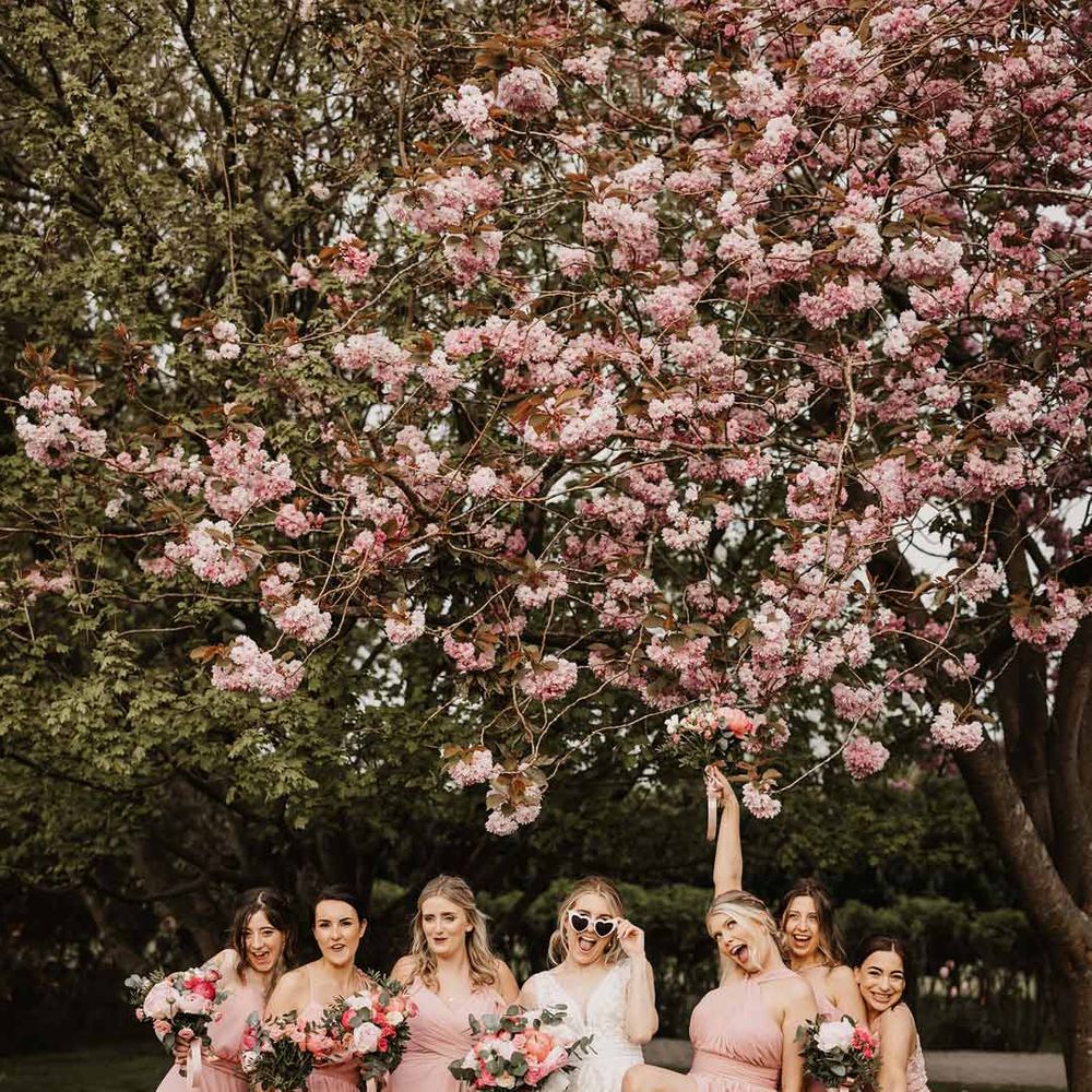 Bride in off shoulder lace wedding dress posing with bridesmaids in mismatched baby pink bridesmaid dresses underneath pink cherry blossom tree