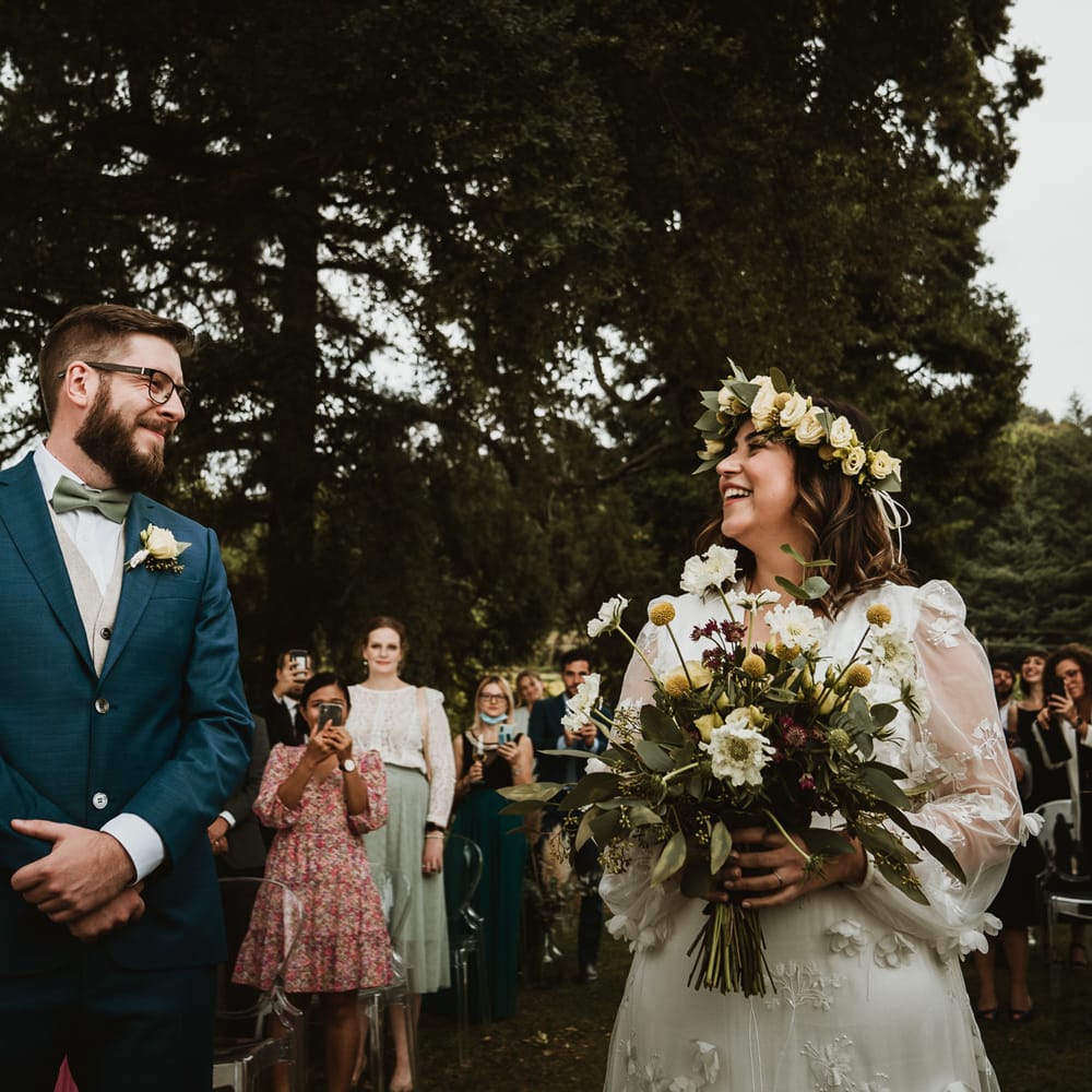 Bride in a long sleeve wedding dress with appliqué detail smiling at her groom in a blue suit and bow tie at the altar 