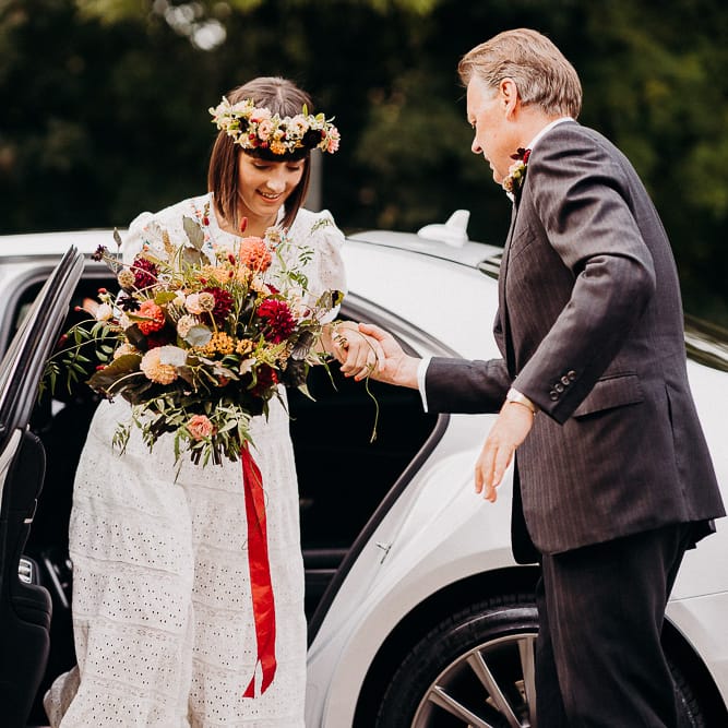 Bride getting our of the white wedding car in a white Broderie Anglaise wedding dress with colourful flower crown and bouquet 