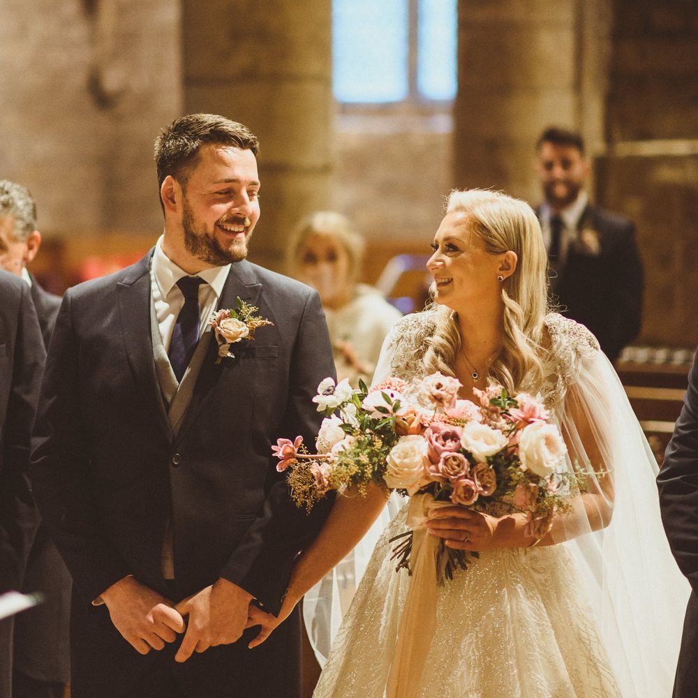 Bride and groom smiling at the altar with bride in a wedding cape holding a blush bouquet 