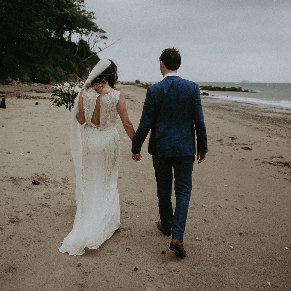 Bride & groom walk hand in hand along the beach on their wedding day