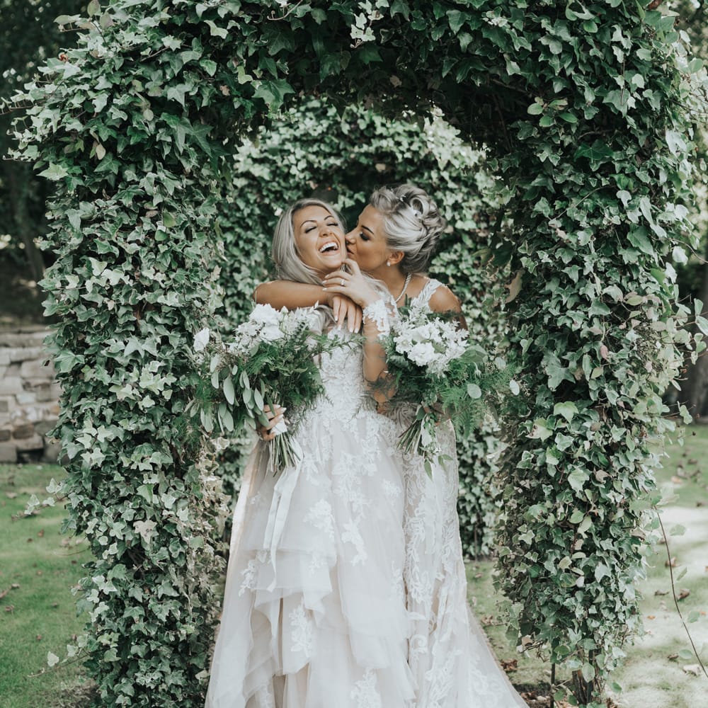 Two brides embrace under ivy covered arches at Crab & Lobster fairytale wedding.