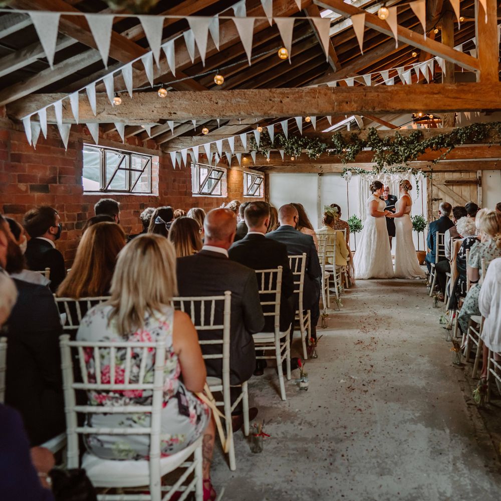Rustic wedding venue complete with white bunting as brides stand at the front of the aisle holding hands during ceremony