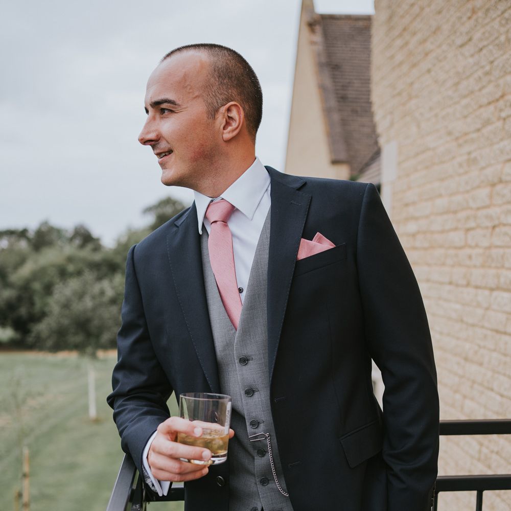 Groom in dark three piece suit with grey waistcoat, pink tie and pink pocket square holding drink whilst he looks out from balcony before Tythe Barn wedding with barn wedding flowers