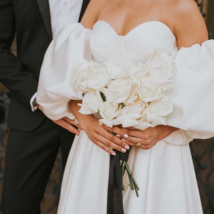 Bride in a sweetheart neckline wedding dress with detachable puff sleeves holding an all white rose wedding bouquet showing off her iridescent nail polish 