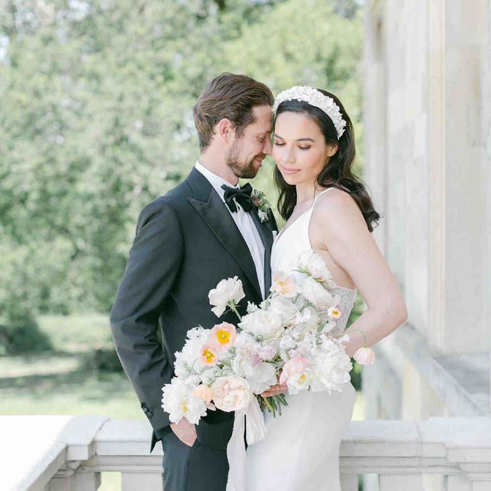 Groom in a tuxedo and bride in an appliqué headband holding a summer wedding bouquet with roses, peonies, icelandic poppies, sweet peas, scabious, foxglove, and snapdragons