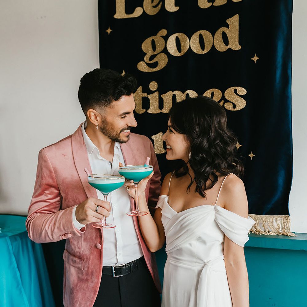 Groom in pink velvet jacket and bride in a cold shoulder wedding dress sipping cocktails in front of a 'let the good times roll' fabric banner 