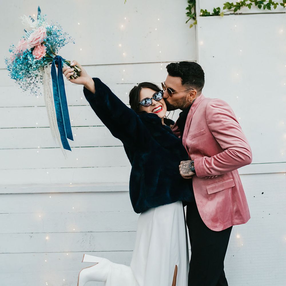 Groom in a pink velvet jacket kissing his brides cheek in a navy faux fur coat as she holds her pink and teal wedding bouquet in the air 