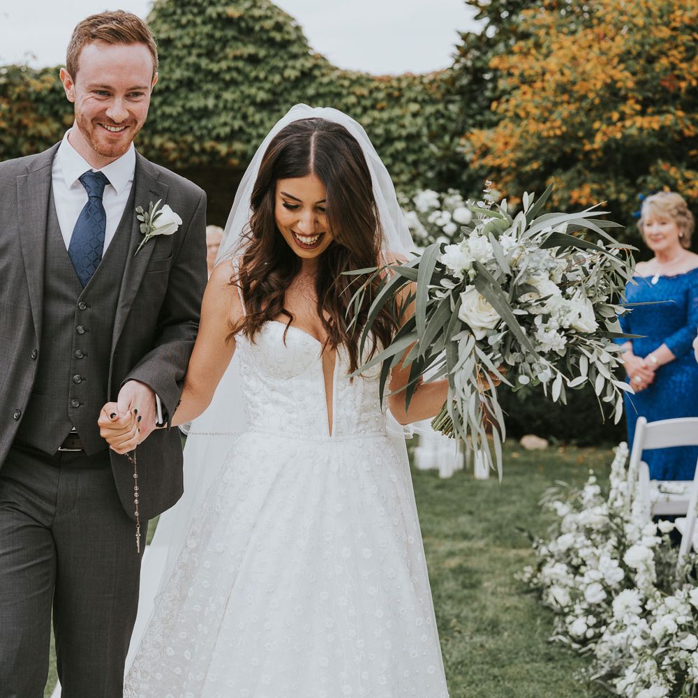 Bride in white cami strap applique Pronovias wedding dress and veil holding green and white rose bridal bouquet walks down the aisle holding hands with groom in grey three piece suits with blue tie and white rose buttonhole during outdoor wedding ceremony in Buckinghamshire