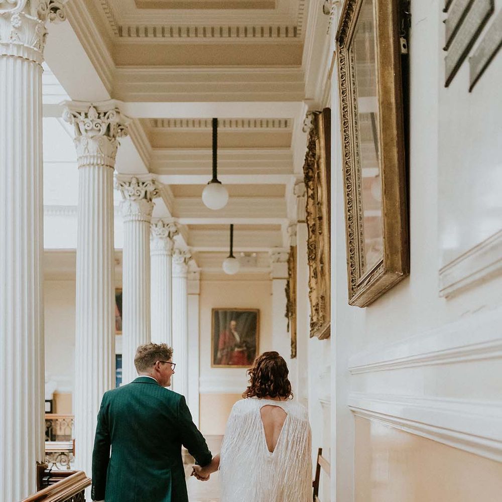 Bride & groom walk away together whilst holding hands and bride wears tassel wedding cape 
