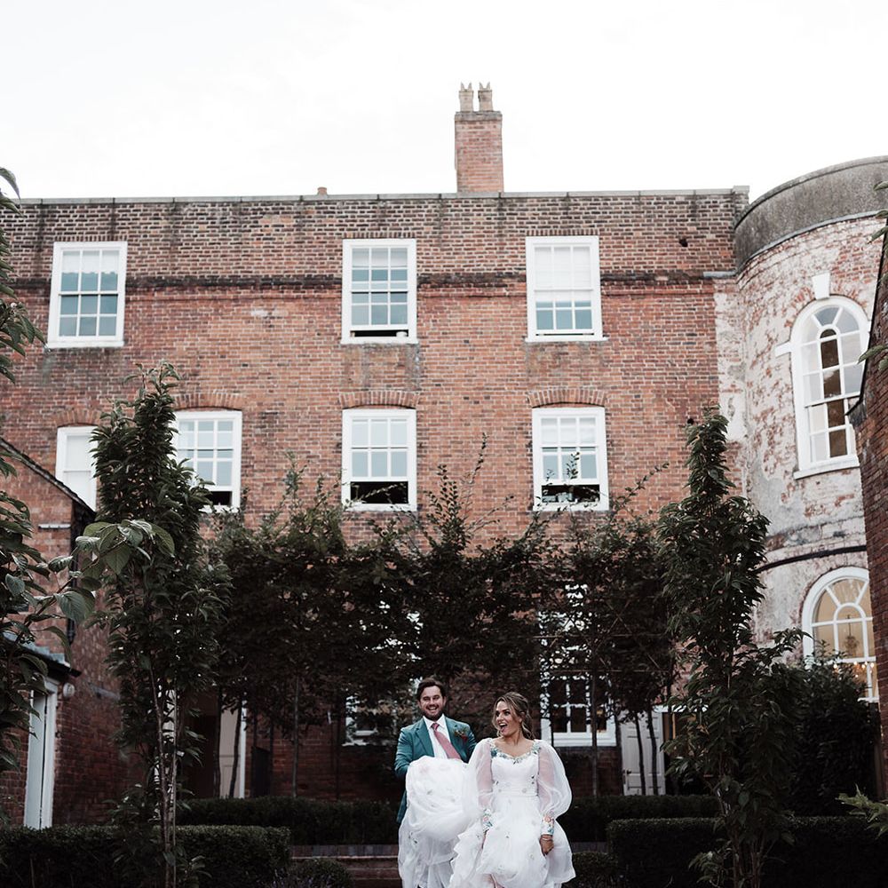 Bride in long sleeve wedding dress and white trainers walks through gardens whilst groom in teal suit holds her train
