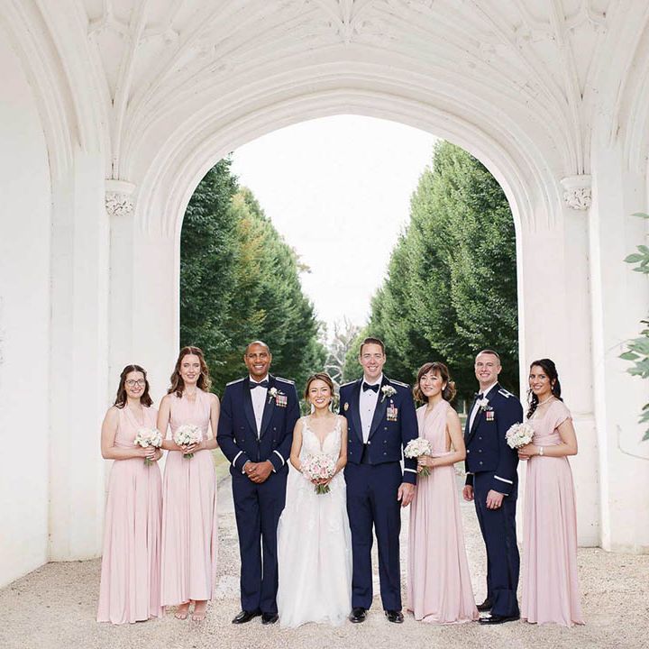 Bride & groom stand with their bridesmaids and groomsmen outdoors under large archway 