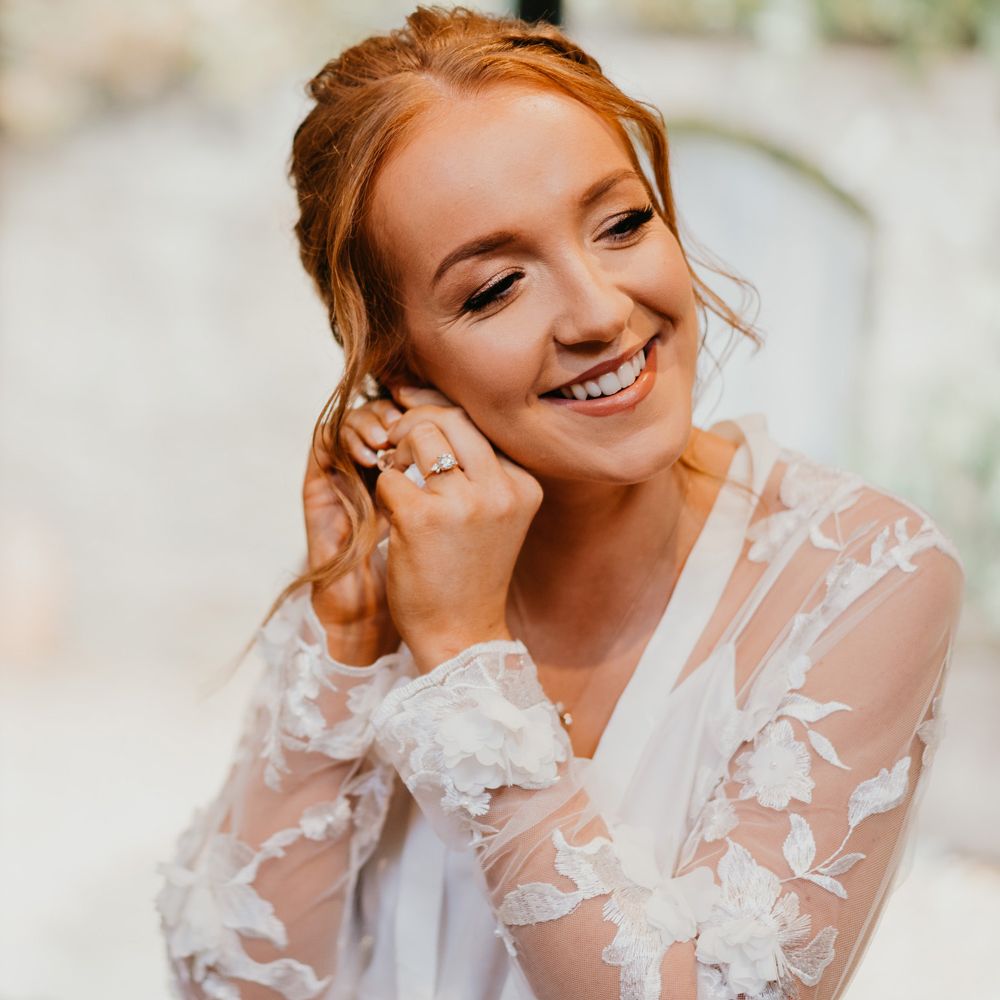Bride with curled updo waiting mesh and lace bridal robe puts in earrings before her wedding