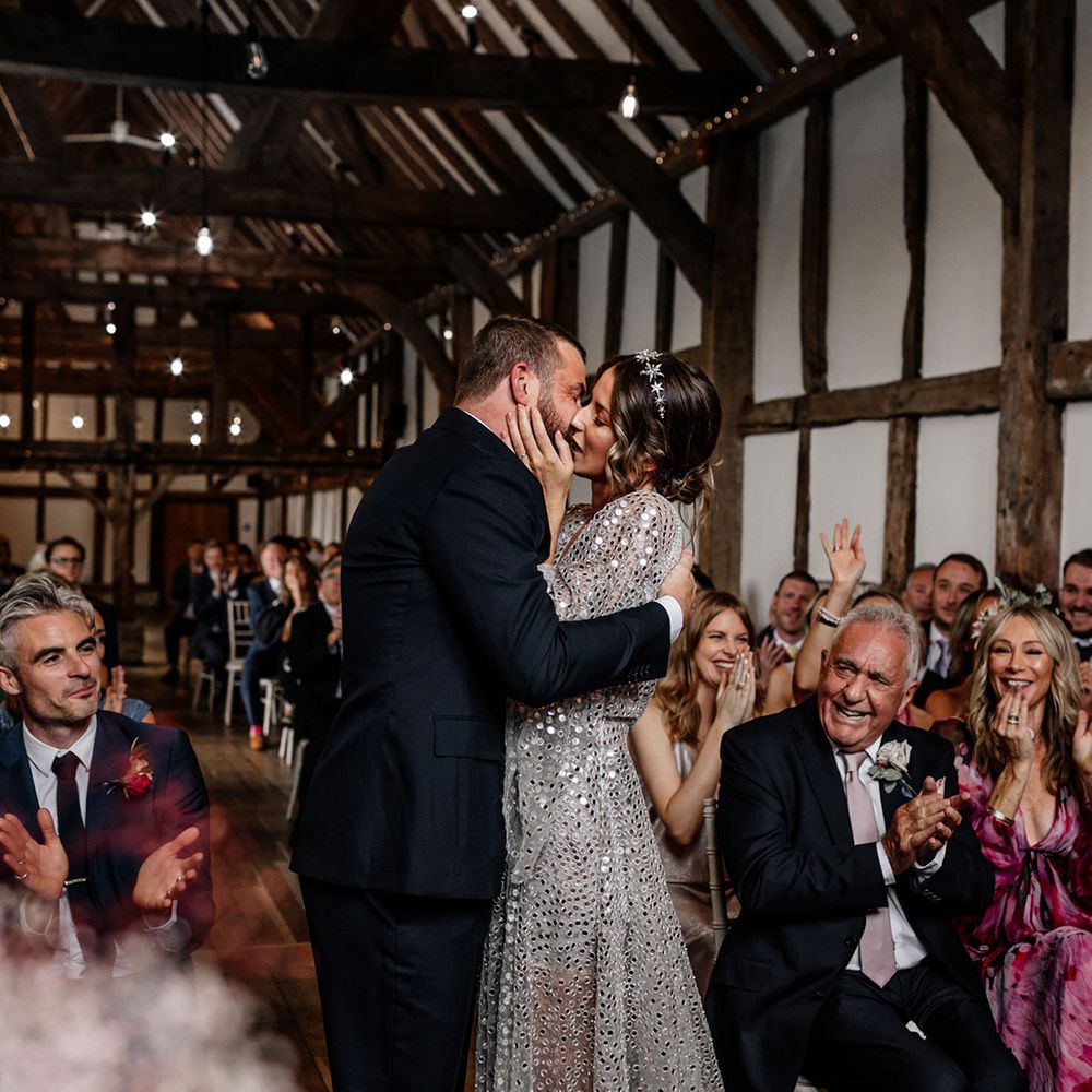 Bride in a silver sequin wedding dress and groom in a navy suit kissing at their Tithe Barn, Loseley House wedding ceremony 