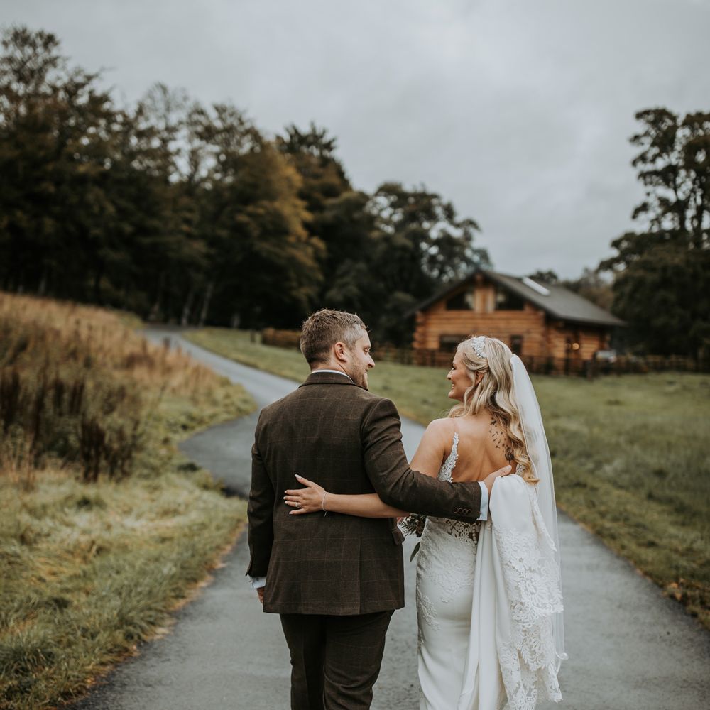 Bride in low back lace wedding dress with button detailing and train walks arm in arm with groom in brown suit as they walk along path at the Hidden River Cabins