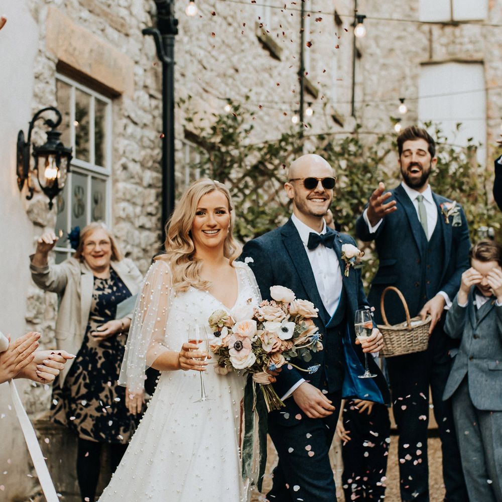 Bride in pearl wedding dress with sleeves holding pastel wedding bouquet and glass of sparkling wine walks out of ceremony with groom navy tweed wedding suit and sunglasses as guests throw confetti