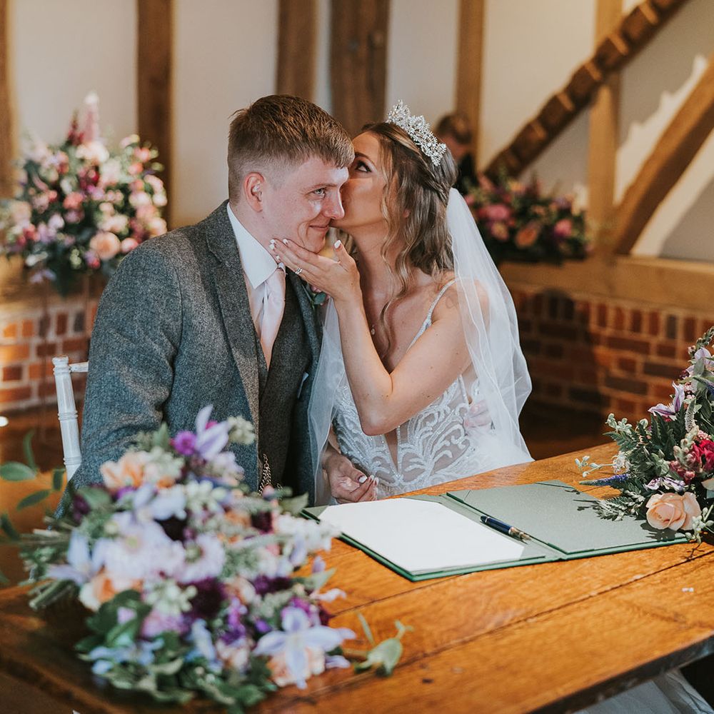 Bride and groom in grey tweed suit sign the registry in rustic wedding venue and large flower displays