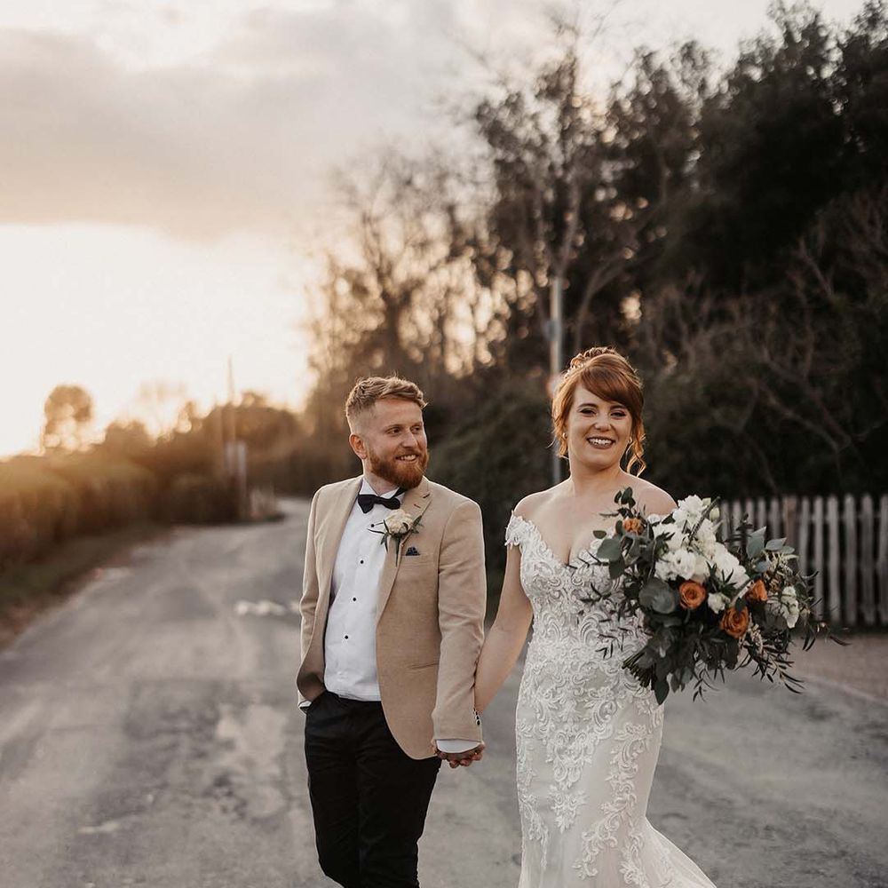 Bride in off the shoulder lace wedding dress with train and groom in beige coloured suit jacket