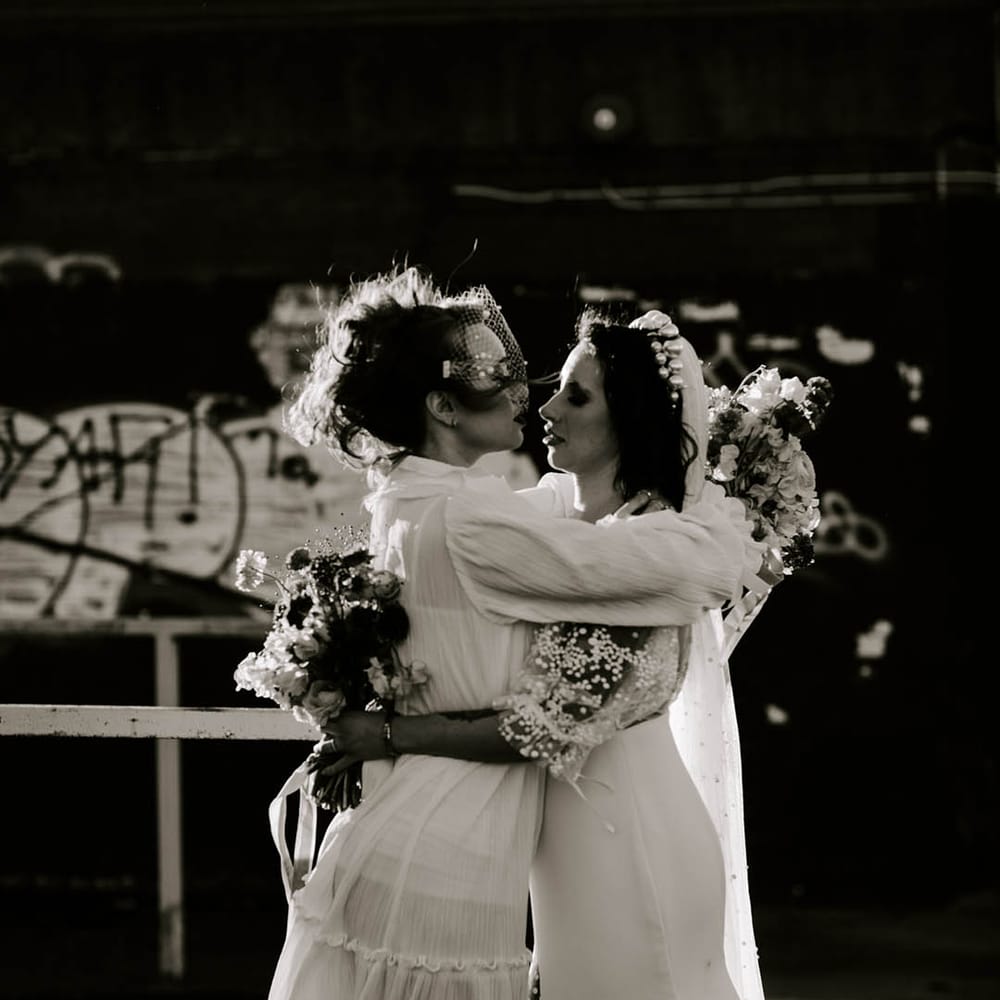 Two brides embracing in long and short wedding dress with a birdcage veil and long embellished veil 