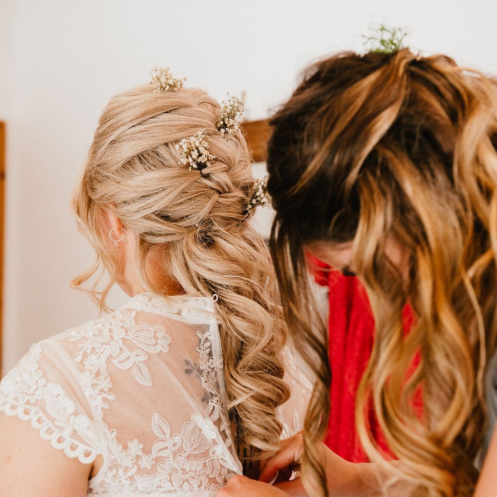 Bride with blonde hair styled into a delicate braid with gypsophila hair pins is buttoned into her wedding dress by friend