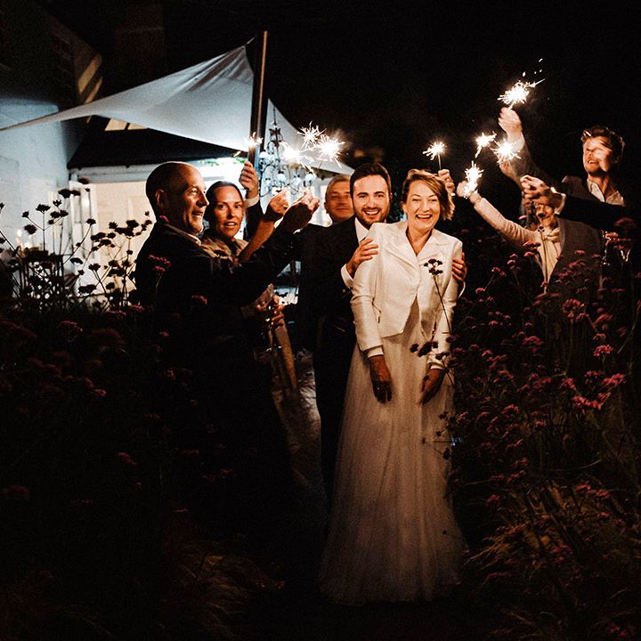 Groom leads the bride wearing white leather cover up jacket through guests waving sparklers