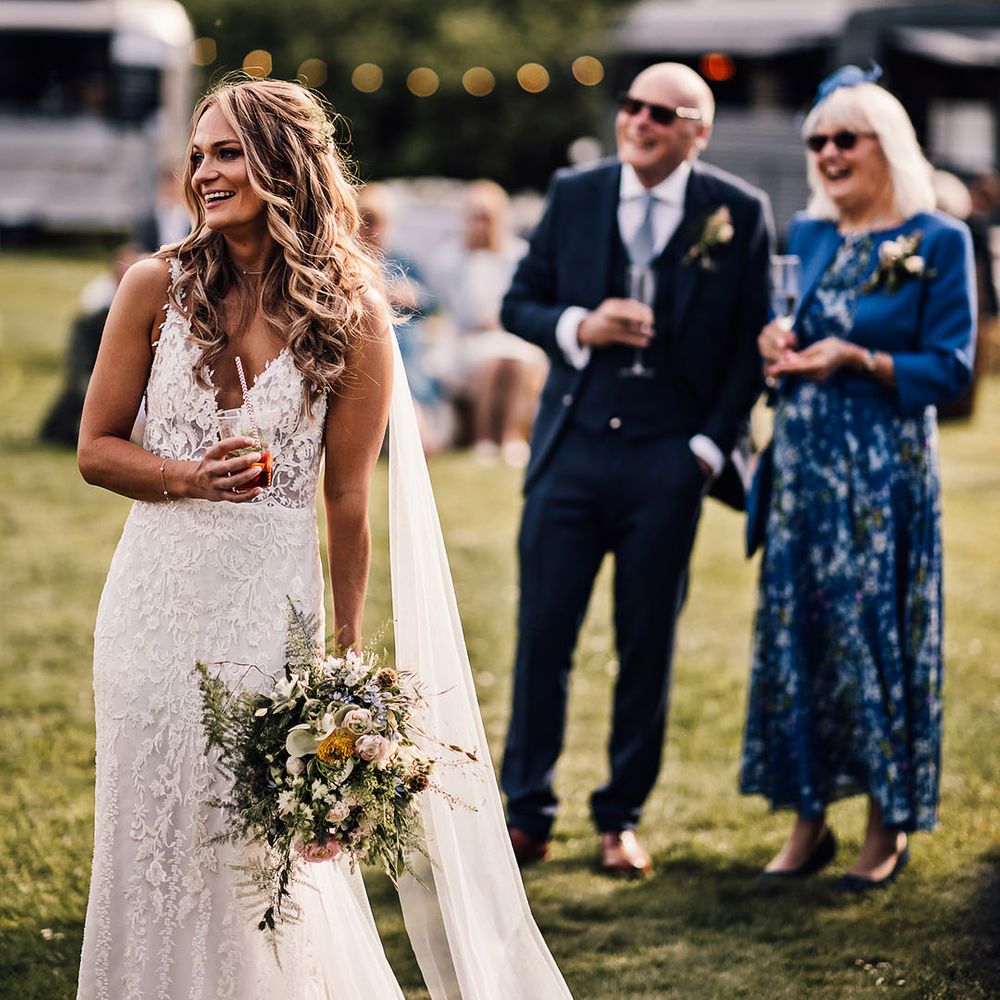 Bride smiles as she listens to the wedding speeches holding a plastic cup and paper straw drink 