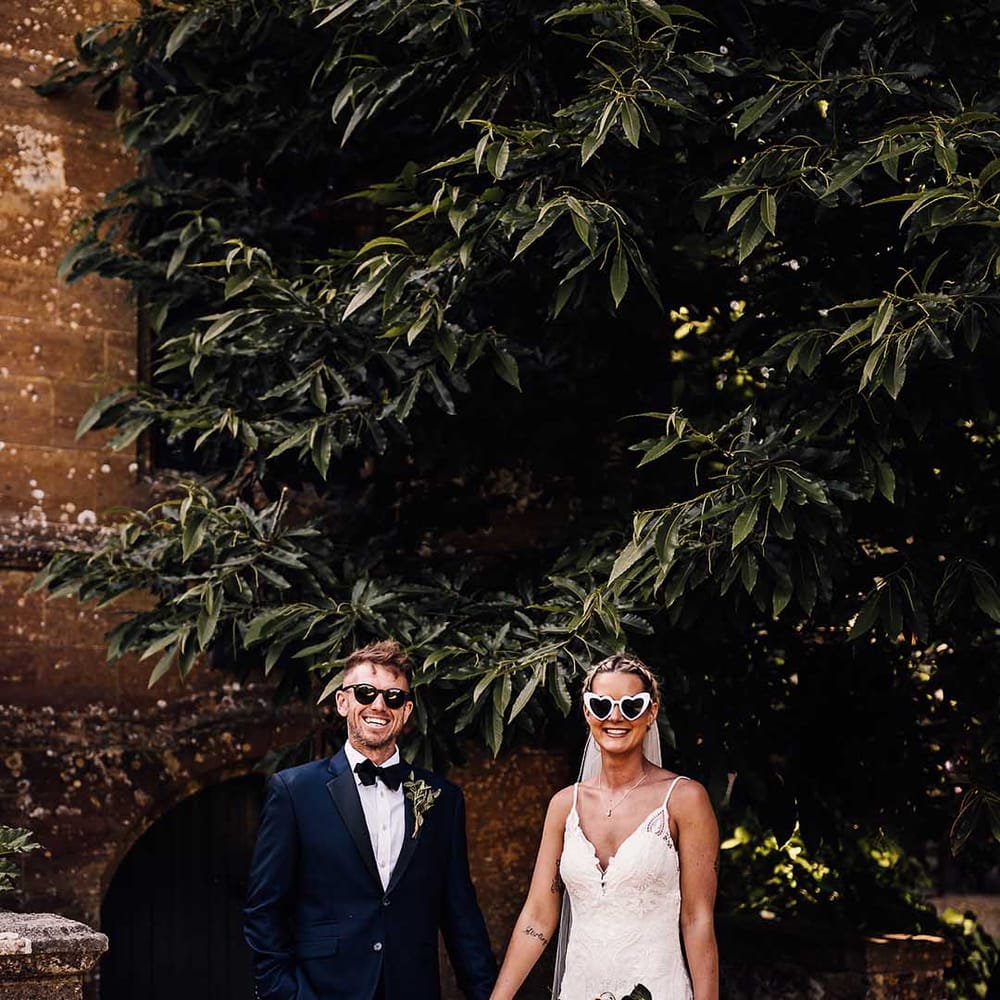 Groom in blue tuxedo and black sunglasses with bride in lace wedding dress and retro heart sunglasses 