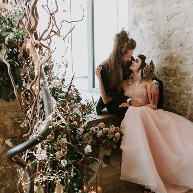 Bride in a pink strapless wedding dress sitting on the steps at Hooton Pagnell Tithe Barn with her groom in a black tuxedo 