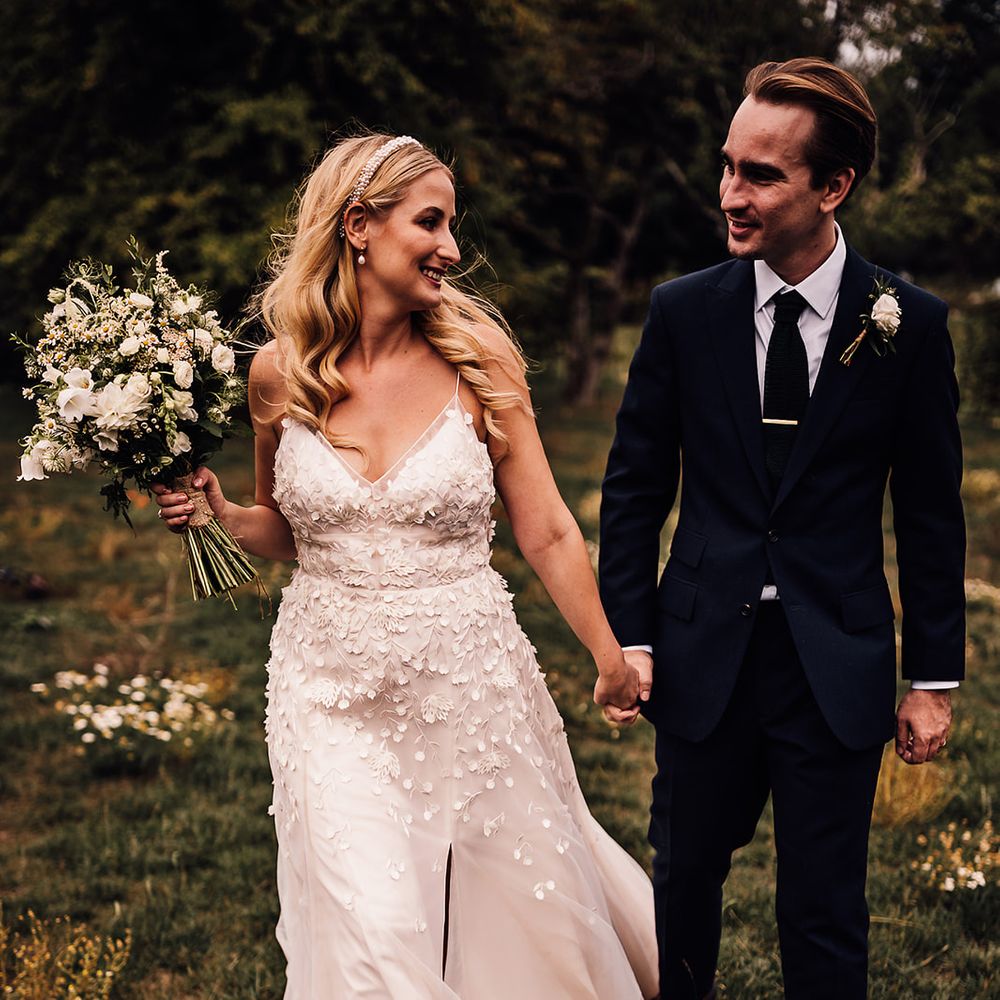 Bride wearing a pearl headband and pearl earrings in an applique wedding dress walks with the groom in a navy blue suit 