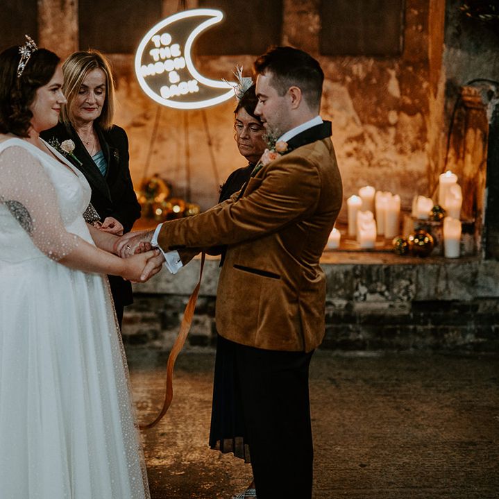 Bride and Groom take part in handfasting ceremony surrounded by candles and a moon neon sign for wedding lighting