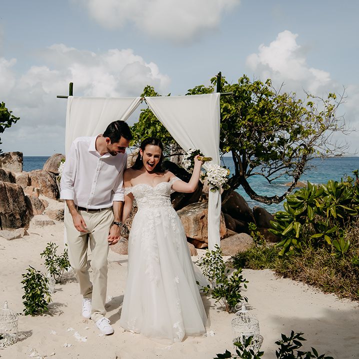 Groom in crisp white shirt, light coloured chinos, tan leather belt and white trainers holding hands with bride in off the shoulder lace wedding dress with sweetheart neckline and lace floral details and cathedral length veil holding white tropical bouquet 