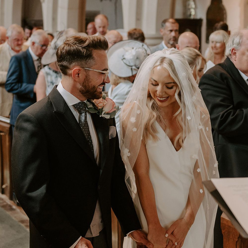 The bride wearing a flower applique veil and satin wedding dress smiling with the groom in three piece suit with large buttonhole for the church ceremony