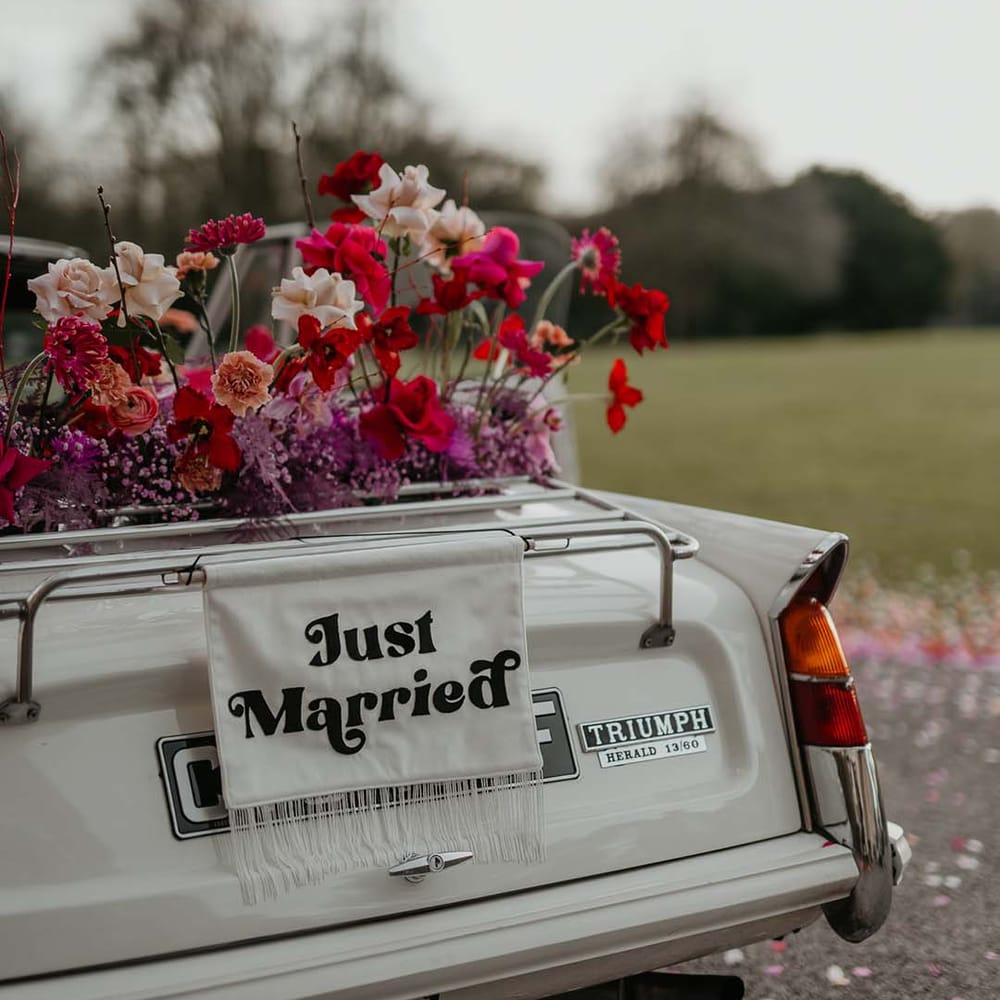 Off white classic convertible wedding car with red roses, off white peonies, lilac dried lowers and white fabric “just married”  banner decorations