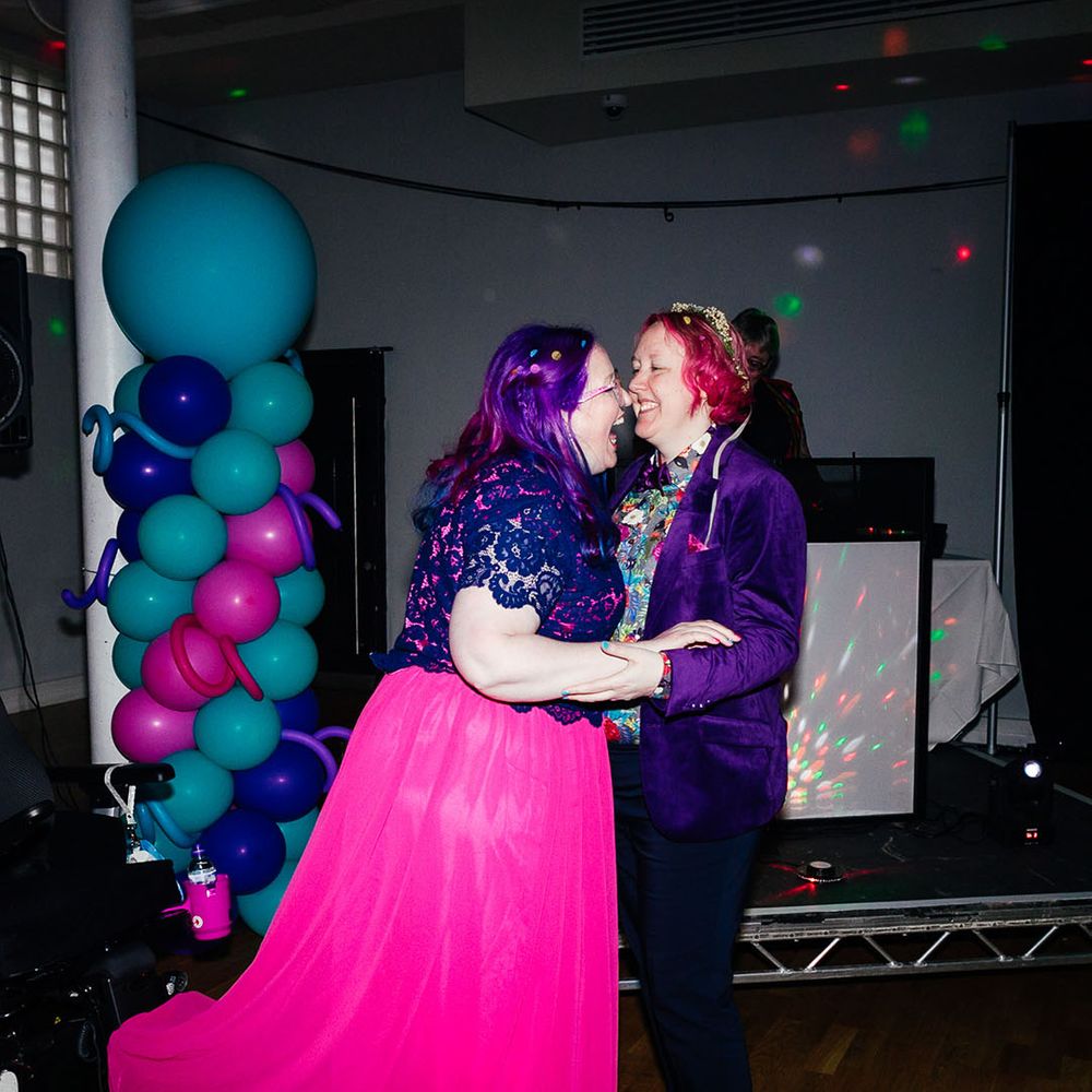 Bride in long pink wedding dress with purple sparkly bridal cover up embracing bride in purple velvet blazer and black suit trousers with purple bowtie, pink patterned pocket square and colourful patterned shirt