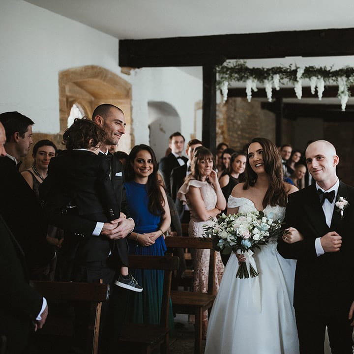 Bride in Pronovias wedding dress walking down the aisle with her brother in black tie 