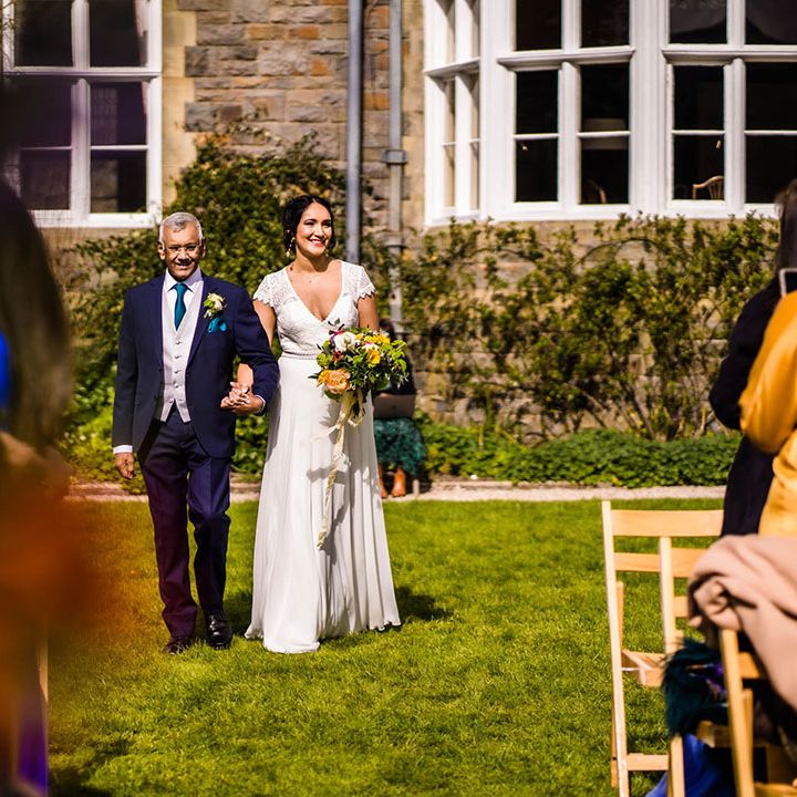 Bride wears lace wedding dress and holds colourful bouquet as she walks down the aisle for outdoor wedding ceremony 