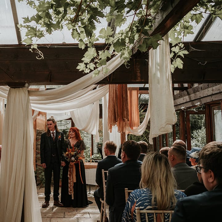 Groom in dark grey three piece suit and burgundy flower patterned tie and bride in embellished black wedding dress holding large orange dried flower bouquet standing amidst off white, white and orange wedding ceiling drapes decor 