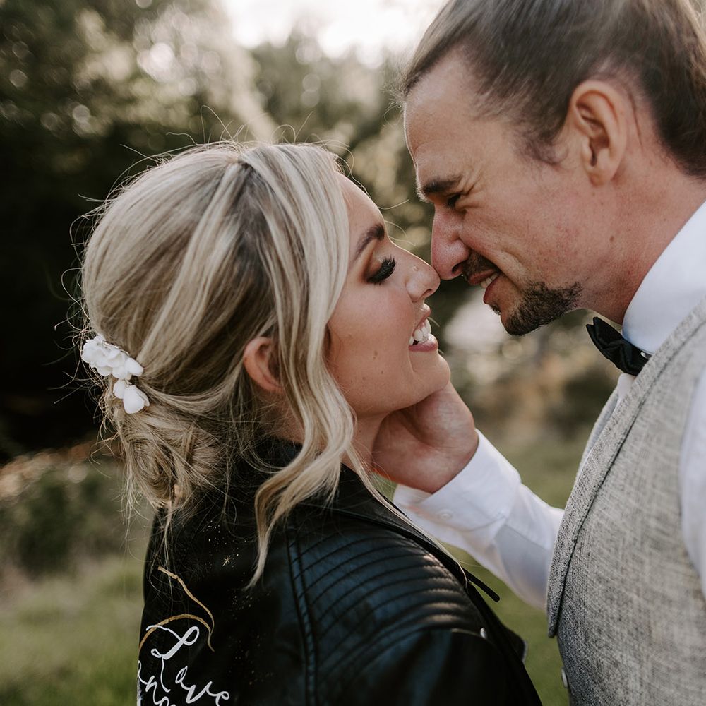 The groom in a light grey waistcoat leans in to kiss the bride who has her hair in an elegant updo in a personalised custom leather jacket 