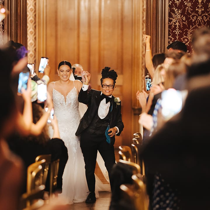 Bride in glasses, black snakeskin print tuxedo with teal lining, black waistcoat, black bowtie and white rose boutonniere walking into the reception room of Grittleton House with bride in lace sleeveless v neck Enzoani wedding dress with tulle finish 