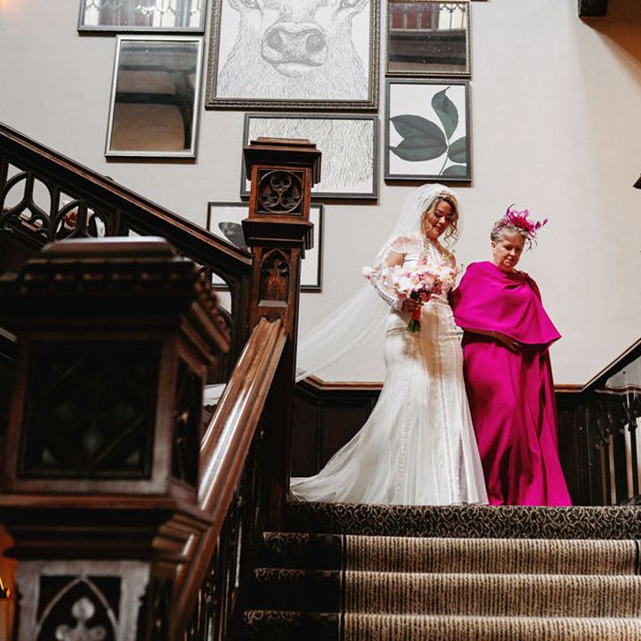 Mother of the bride in bright pink wedding outfit walking down the stairs with the bride in a delicate lace wedding dress 
