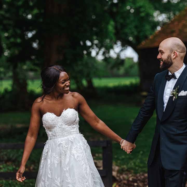 Groom in classic black three piece grooms suit with bow tie and white rose and foliage boutonniere in church holding hands with bride in sweetheart neckline lace wedding dress walking through the grounds after wedding breakfast