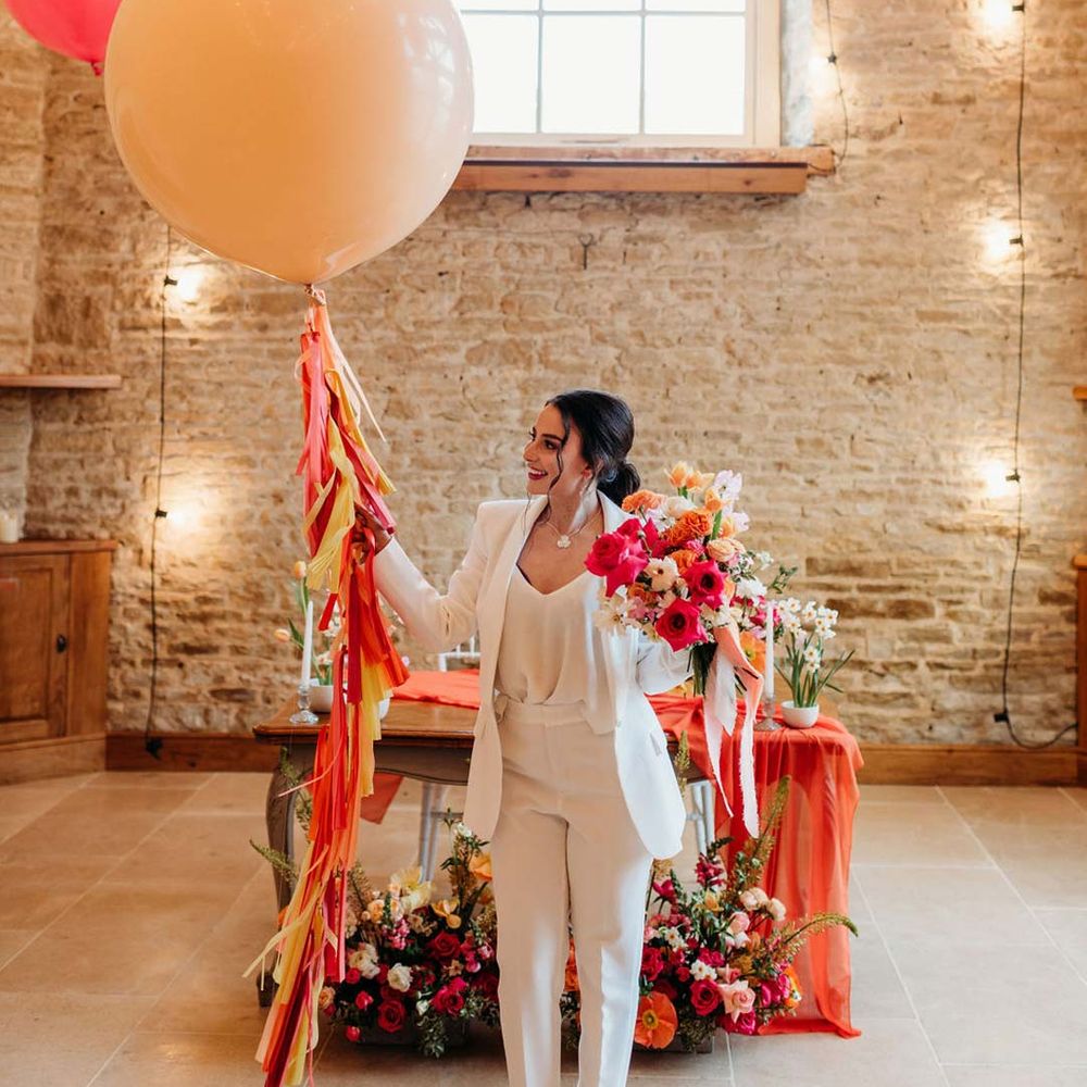 Bride in white bridal suit in Merriscourt wedding venue reception room with metallic pink, white, orange and yellow balloons, wedding fairy lights decorations and wooden table with orange table runner and mixed seasonal flower arrangements 