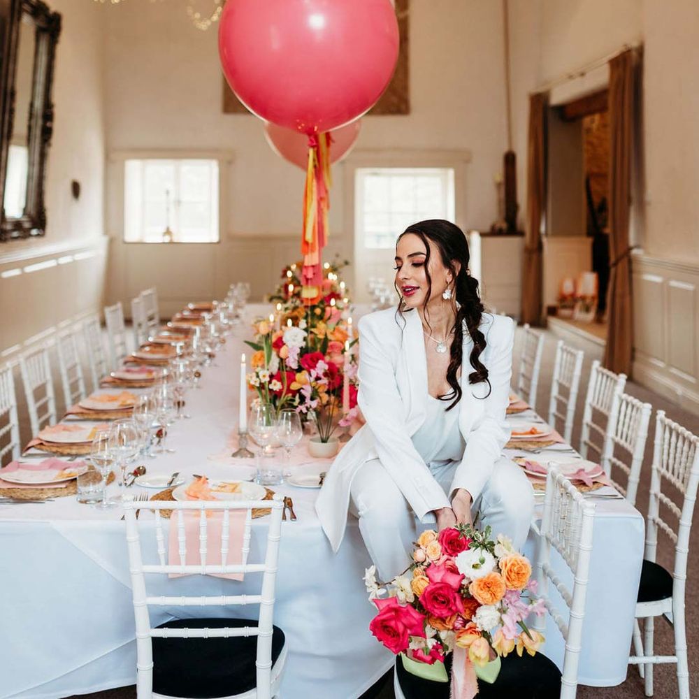 Bride in white bridal suit with silver bridal jewellery with closed toe bright green wedding heels holding colourful bridal bouquet sitting on eclectic colourful wedding tablescape at Merriscourt wedding venue